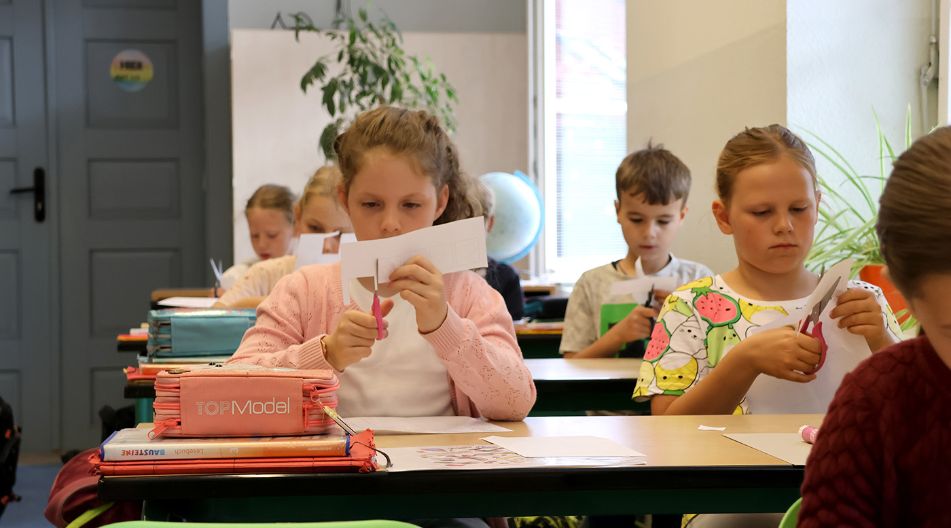 Zwei Schülerinnen sitzen am Schultisch schneiden im Rahmen des Lesebandes konzentriert etwas aus, um es später aufzukleben.