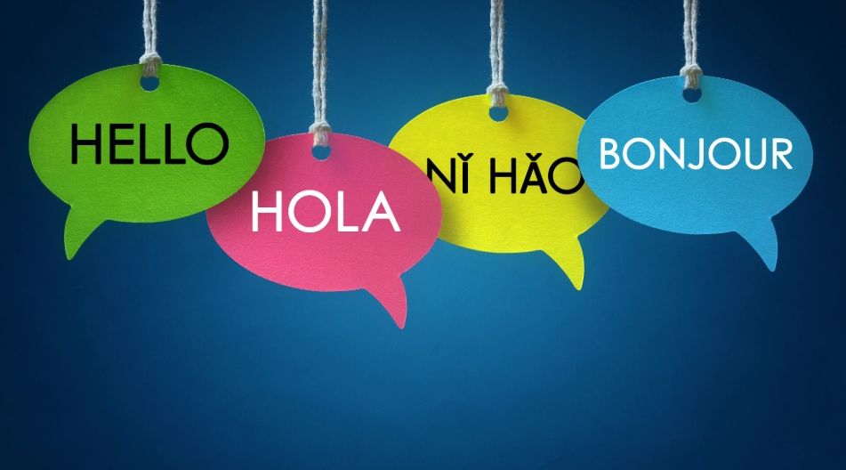 Vier bunte Sprechblasen mit dem Wort „Hallo“ in verschiedenen Sprachen: Englisch (Hello), Spanisch (Hola), Chinesisch (Nǐ hǎo) und Französisch (Bonjour), hängen nebeneinander an Schnüren vor blauem Hintergrund.