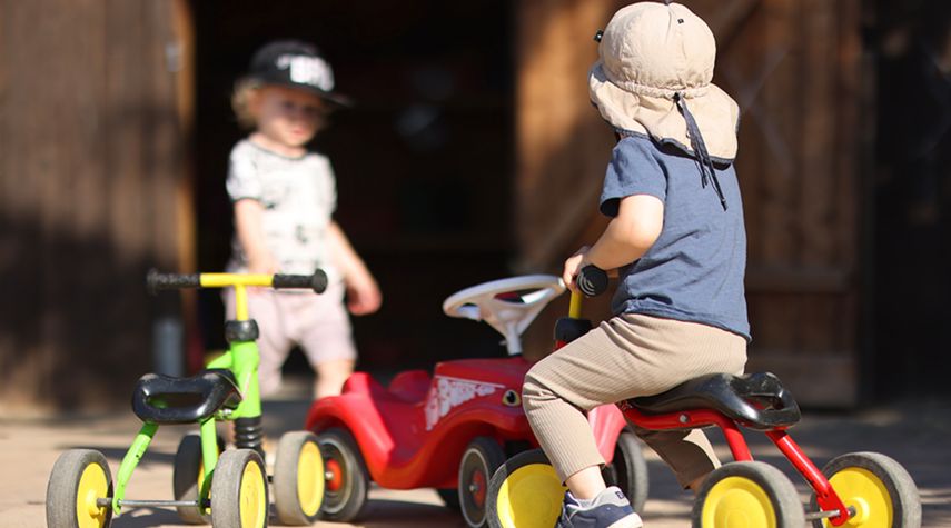 Ein Kind mit Sonnenhut sitzt auf einem roten Dreirad, während ein weiteres Kind mit Baseballkappe im Hintergrund steht. Mehrere bunte Kinderfahrzeuge stehen auf einem sonnigen Hof. Ein Kind mit Sonnenhut sitzt auf einem roten Dreirad, während ein weiteres Kind mit Baseballkappe im Hintergrund steht. Mehrere bunte Kinderfahrzeuge stehen auf einem sonnigen Hof.