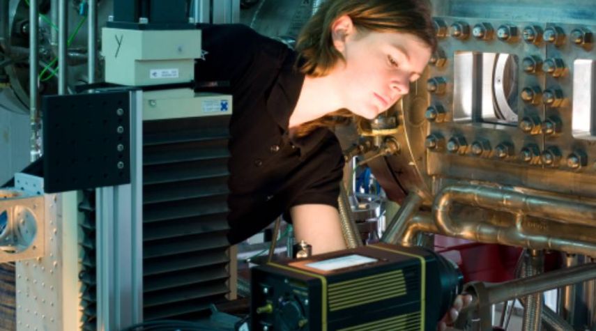 Eine junge Frau in schwarzer Arbeitskleidung arbeitet konzentriert an einer technischen Apparatur mit Metallrohren und Messgeräten in einem Labor oder einer industriellen Umgebung.