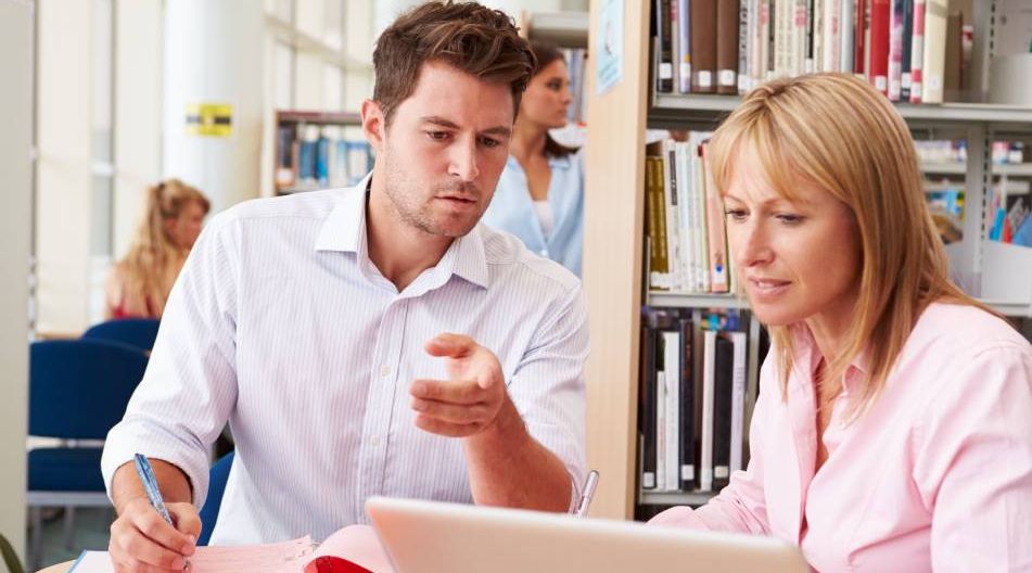 Zwei Erwachsene arbeiten gemeinsam in einer Bibliothek an einem Laptop, umgeben von Büchern.