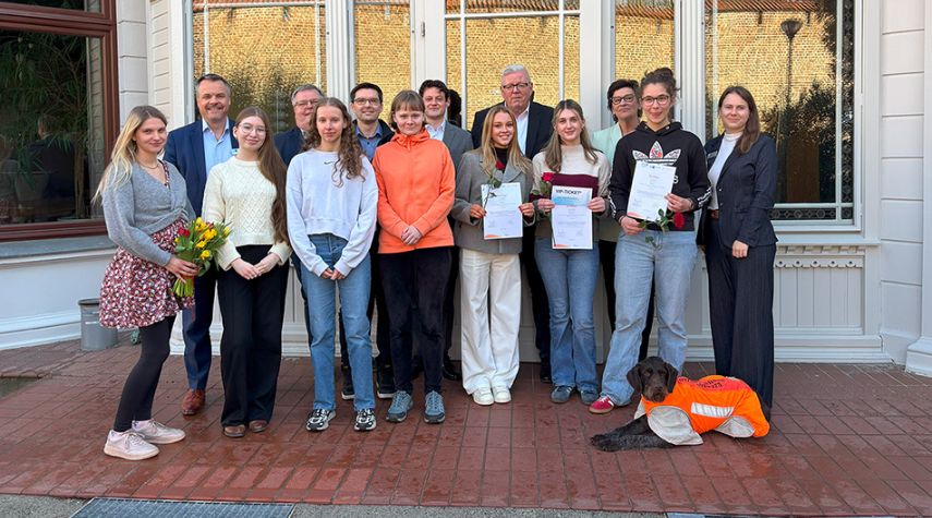 Gruppe von Jugendlichen und Erwachsenen vor einem Gebäude, einige halten Urkunden und Blumen; ein Hund mit Warnweste liegt vorne.