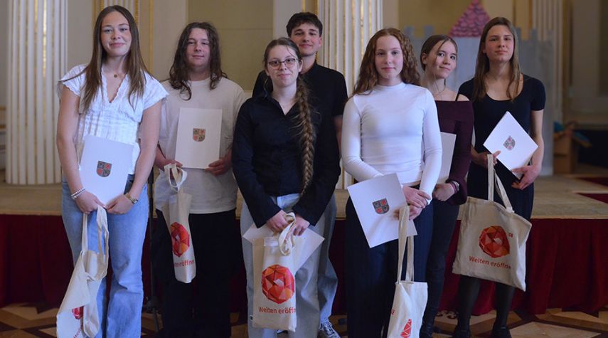 Gruppe von sieben Jugendlichen steht in einem festlichen Saal und hält Urkunden sowie Stofftaschen nach der Preisverleihung beim Fremdsprachenwettbewerb.