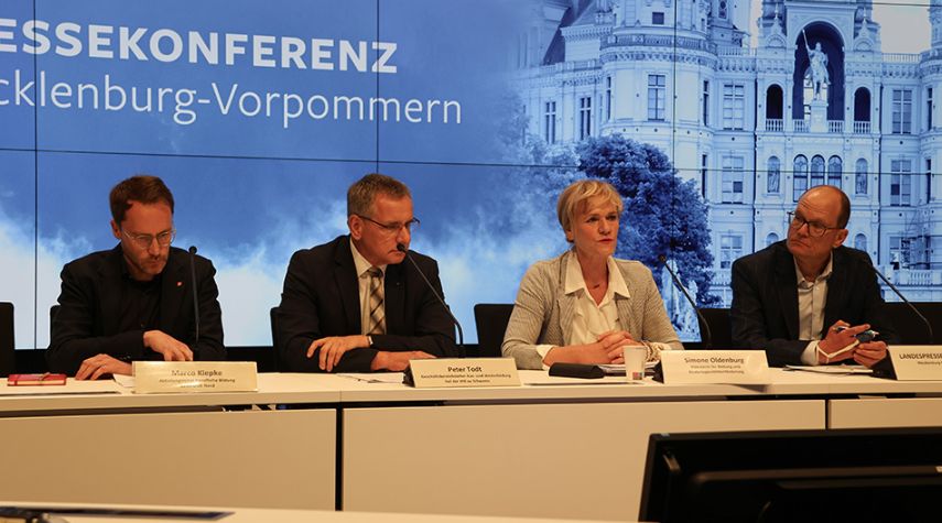 Vier Personen sitzen bei einer Pressekonferenz in Mecklenburg-Vorpommern an einem Tisch mit Mikrofonen, vor einer großen Präsentationswand.