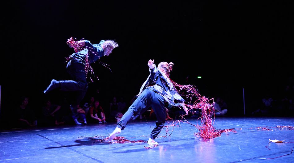 Zwei Tänzerinnen bewegen sich dynamisch auf einer dunklen Bühne, rote Bänder wirbeln im Bühnenlicht um sie herum, Publikum sitzt im Hintergrund.