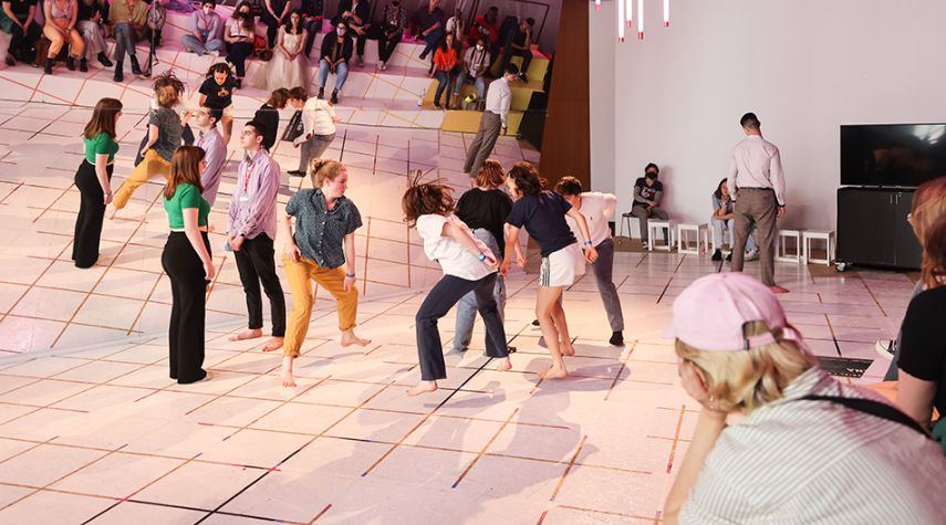 Mehrere Personen tanzen barfuß auf einer Bühne, während das Publikum auf Stufen im Hintergrund zuschaut.