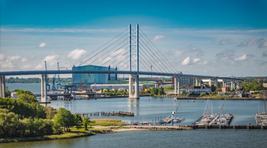 Blick auf die Rügenbrücke über das Wasser mit Hafen, Booten und Industrieanlagen im Hintergrund bei klarem Himmel.