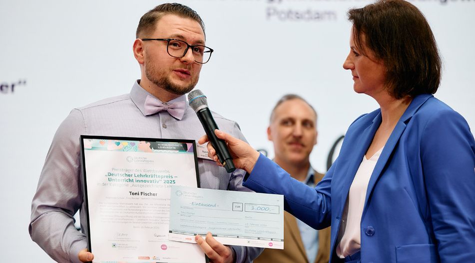 Ein Mann mit Brille und Fliege hält eine gerahmte Urkunde und einen Scheck, während eine Frau im blauen Blazer ihm bei einer Preisverleihung ein Mikrofon hinhält.