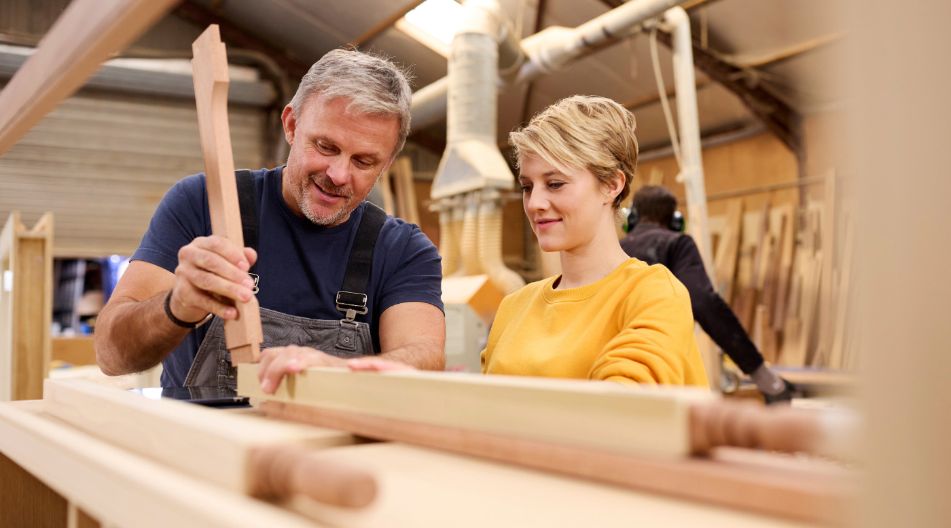 Ein erfahrener Schreiner zeigt einer jungen Frau in einer Werkstatt den Umgang mit einem Holzwerkstück, während sie aufmerksam zusieht.