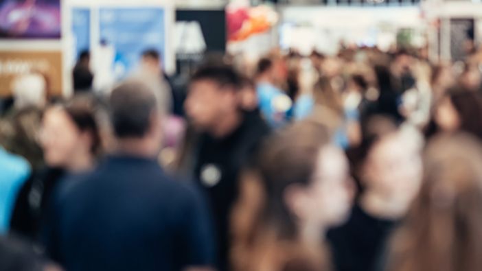 Eine verschwommene Aufnahme von einer Berufsmesse. Viele Menschen gehen einen Gang entlang oder stehen an den Seiten bei den verschiedenen Ständen.