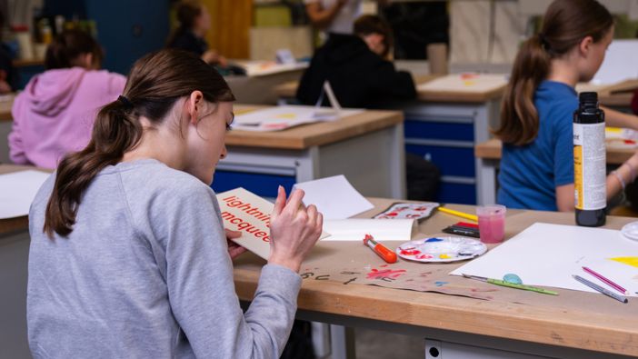 Schülerinnen und Schüler arbeiten in einem Klassenzimmer an kreativen Projekten, mit Bastelmaterialien wie Farben, Pinseln und Papier auf den Tischen."