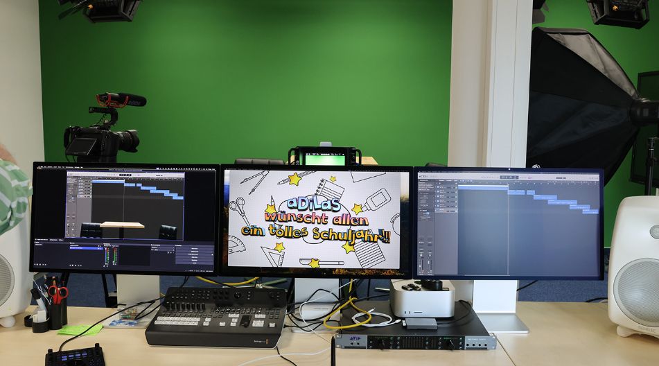 Ein modernes Video- und Tonaufnahmestudio mit drei Monitoren, die eine Videoproduktion und Tonbearbeitung zeigen. Im Hintergrund befindet sich eine grüne Wand (Greenscreen) sowie Kamera- und Beleuchtungsausrüstung. Auf dem mittleren Monitor steht „aDilas wünscht allen ein tolles Schuljahr!!“ in bunten Buchstaben.