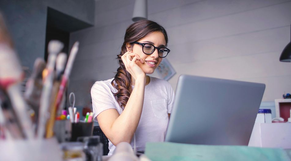 Junge Frau mit Brille sitzt an einem Schreibtisch, schaut lächelnd auf einen Laptop und ist von Bastel- und Büromaterialien umgeben.