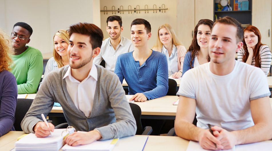 Eine Gruppe junger Erwachsener sitzt in einem Klassenraum, hört aufmerksam zu und macht sich Notizen. Sie wirken motiviert und konzentriert.