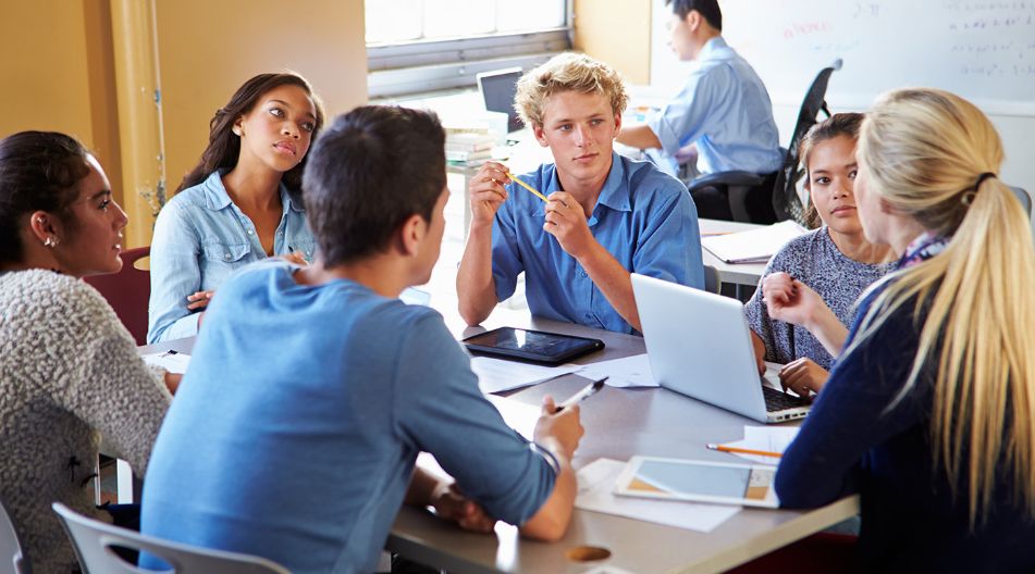 Eine Gruppe von sechs jungen Erwachsenen sitzt an einem Tisch in einem Klassenzimmer und diskutiert. Sie sind von Laptops, Tablets, Notizbüchern und Stiften umgeben. Im Hintergrund arbeitet eine weitere Person an einem Schreibtisch mit einem Laptop.