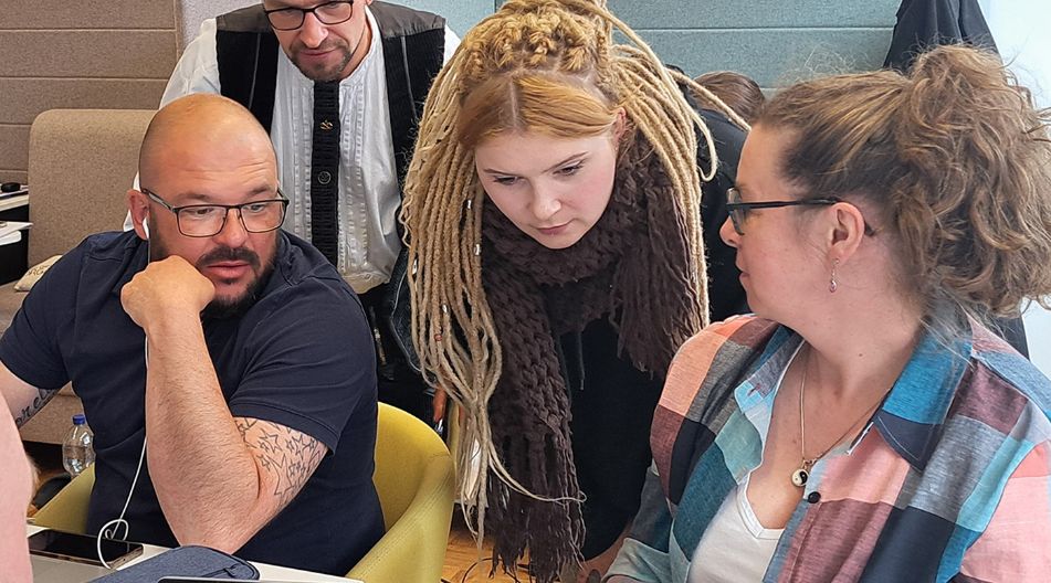 Vier Erwachsene arbeiten gemeinsam an einem Laptop in einem modernen Lernraum. Drei Personen schauen konzentriert auf den Bildschirm, während eine Person stehend über die Schulter blickt. Im Vordergrund ist ein geöffnetes digitales Lernmodul sichtbar.