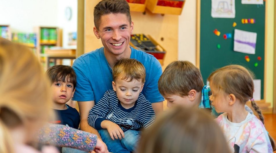 Ein lächelnder Erzieher liest einer Gruppe von Kindern in einem Klassenzimmer ein Buch vor.