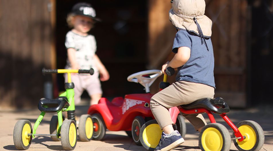 Ein Kind mit Sonnenhut sitzt auf einem roten Dreirad, während ein weiteres Kind mit Baseballkappe im Hintergrund steht. Mehrere bunte Kinderfahrzeuge stehen auf einem sonnigen Hof. Ein Kind mit Sonnenhut sitzt auf einem roten Dreirad, während ein weiteres Kind mit Baseballkappe im Hintergrund steht. Mehrere bunte Kinderfahrzeuge stehen auf einem sonnigen Hof.