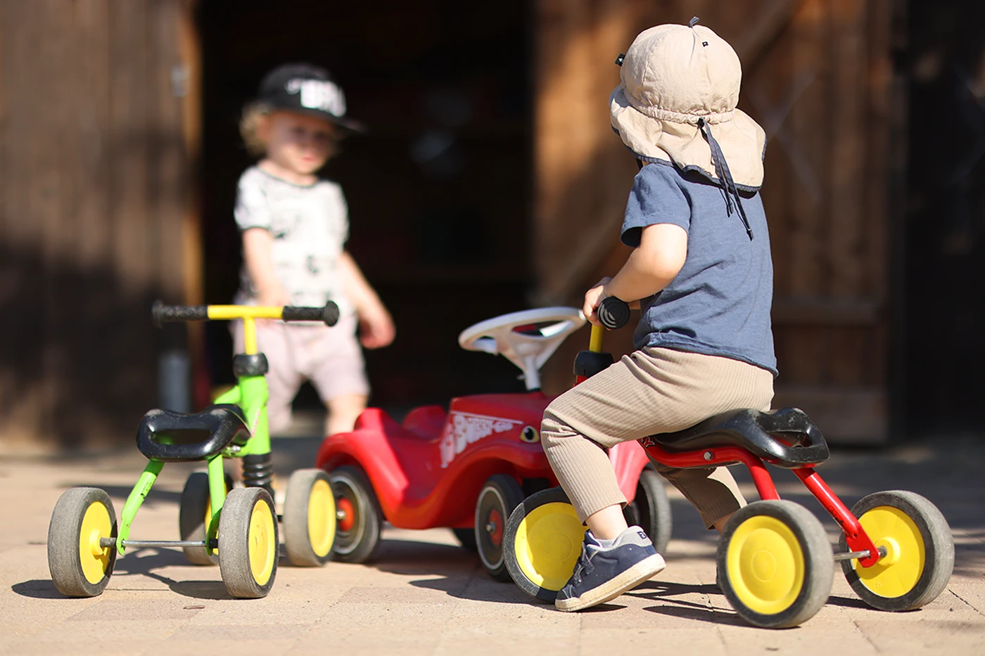 Ein Kind mit Sonnenhut sitzt auf einem roten Dreirad, während ein weiteres Kind mit Baseballkappe im Hintergrund steht. Mehrere bunte Kinderfahrzeuge stehen auf einem sonnigen Hof.