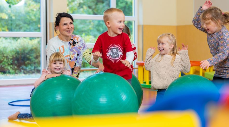 Kinder spielen lachend mit großen Gymnastikbällen, betreut von einer Erzieherin.