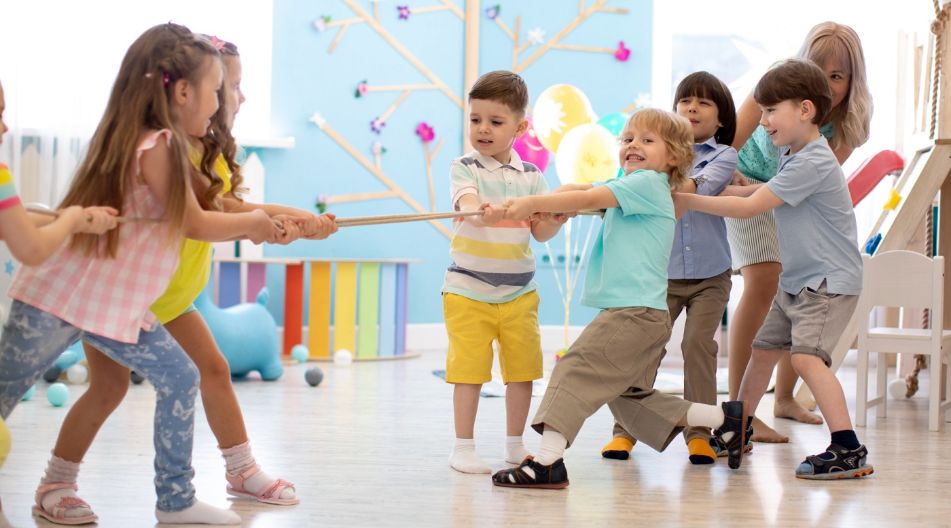 Eine Gruppe von Kindern spielt Tauziehen in einem hellen, farbenfrohen Raum. Die Kinder lächeln und sind in zwei Teams aufgeteilt, die jeweils an einem Seil ziehen, unterstützt von einer Betreuerin im Hintergrund.