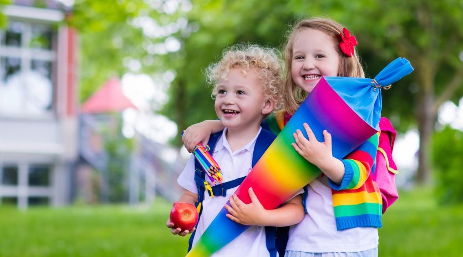 Zwei lachende Kinder mit Schultüte und Rucksäcken vor einer Schule.