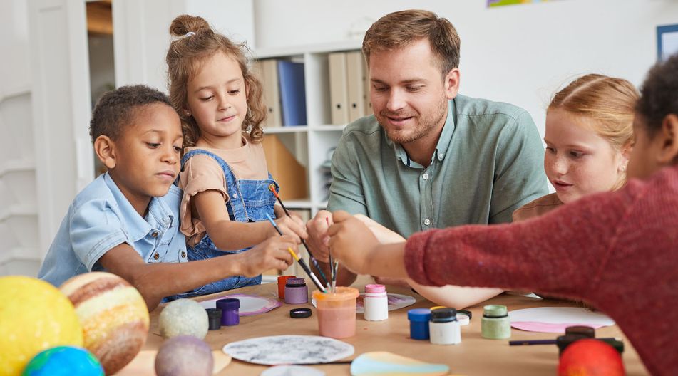 Ein Mann sitzt mit mehreren Kindern an einem Tisch und malt mit ihnen runde Blätter mit Tuschkastenfarbe an.