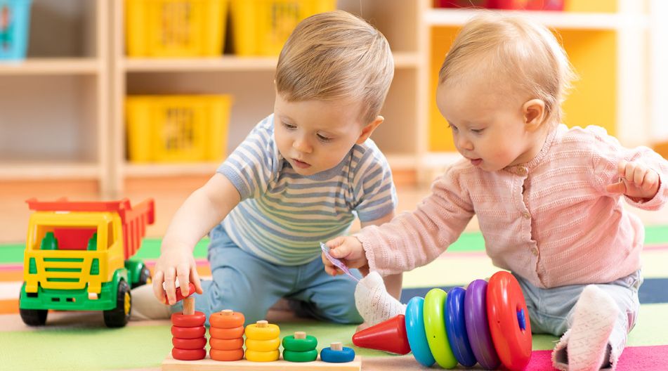 Zwei Kleinkinder spielen auf einer bunten Matte mit Lernspielzeug. Eines trägt ein gestreiftes Shirt und stapelt Ringe auf einem Holzstab, das andere trägt eine rosa Strickjacke und hält ein Plastikteil in der Hand. Im Hintergrund stehen Regale mit gelben Aufbewahrungskörben.