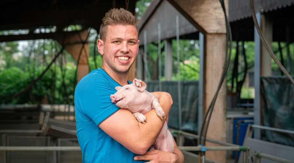 Ein junger Bauer hält vor einem Stall lächelnd ein Schwein im Arm.