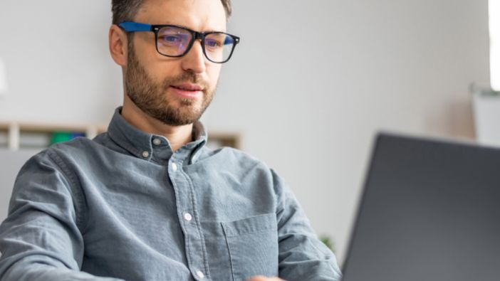 Ein Mann mit blauer Bildschirmarbeitsplatzbrille sitzt an einem Schreibtisch und arbeitet konzentriert an einem Laptop.