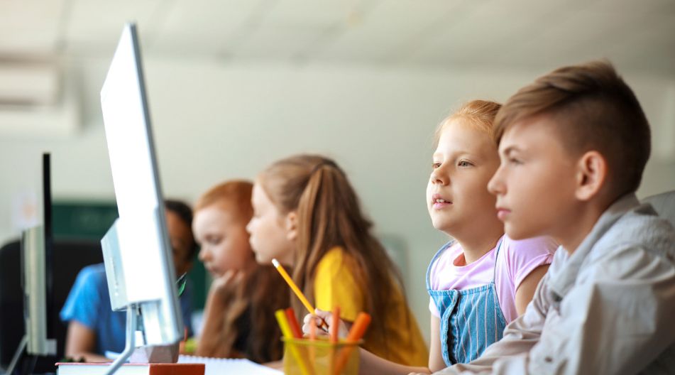 Mehrere Kinder schauen auf einen PC-Monitor und machen sich nebenbei Notizen zu einem Thema.