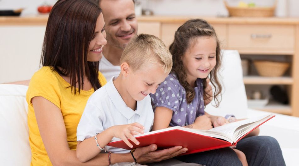 ine Familie sitzt auf dem Sofa und liest gemeinsam in einem großen roten Buch. Die Eltern lächeln, während die beiden Kinder, ein Junge und ein Mädchen, begeistert auf die Seiten schauen.