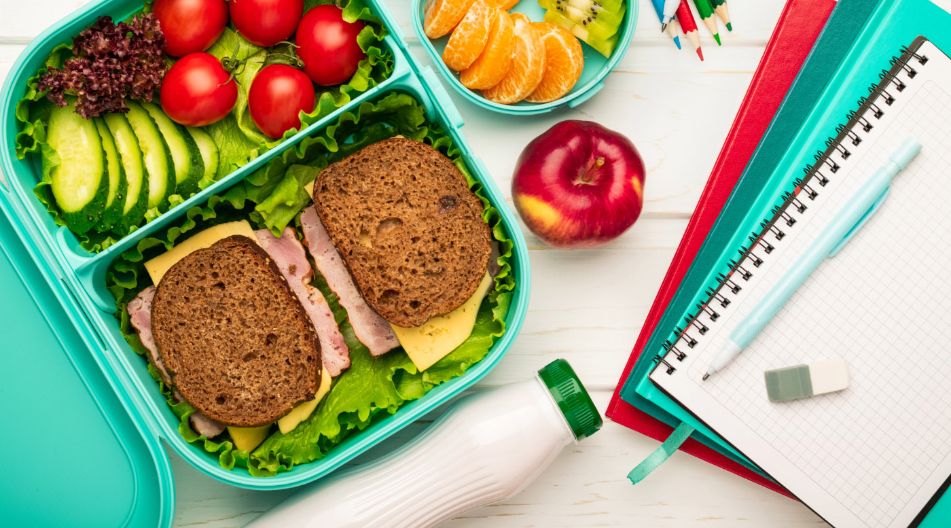 Ein geöffnetes Lunchbox-Set mit Vollkornbrotsandwiches, Tomaten, Gurkenscheiben, Blattsalat, Mandarinenstücken, Kiwi, und einem Apfel. Daneben liegen Schreibutensilien, Hefte und eine Trinkflasche – alles auf einem hellen Holztisch arrangiert.