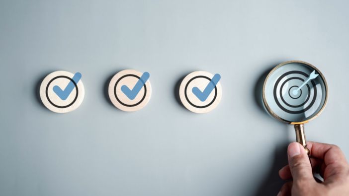 Vier Holzplättchen mit Häkchen liegen nebeneinander, während eine Hand eine Lupe über das letzte Plättchen hält, das ein Zielsymbol mit Pfeil zeigt.