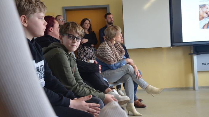 Eine Gruppe von Personen sitzt in einem Klassenraum und hört aufmerksam zu. Im Vordergrund sitzen zwei Jungen, einer davon schaut zur Kamera. Im Hintergrund stehen mehrere Erwachsene vor einer gelben Wand, während rechts ein Bildschirm und eine Tafel zu sehen sind.