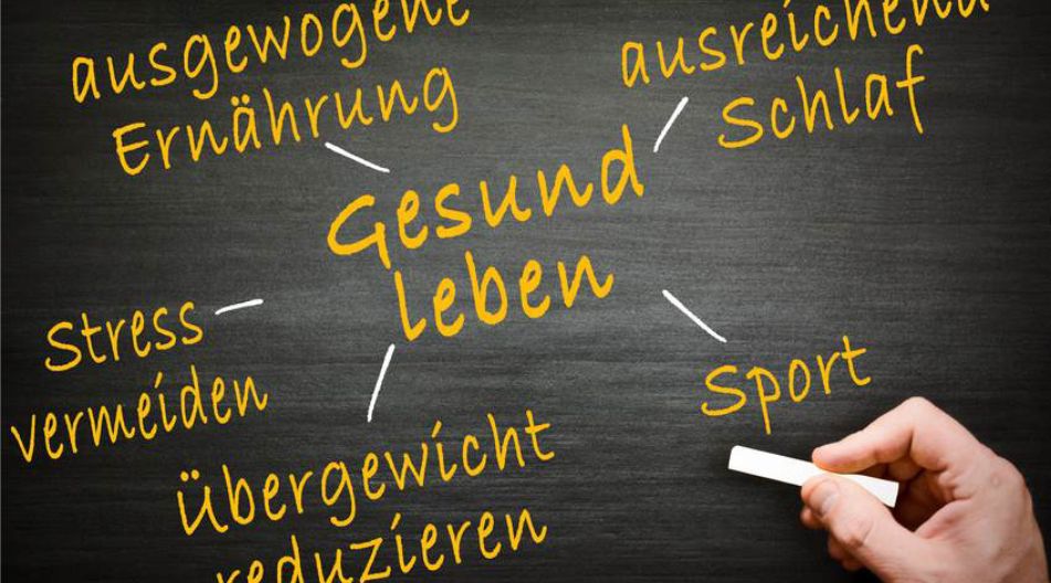 Tafel mit der Aufschrift „Gesund leben“ in der Mitte, umgeben von Begriffen wie „ausgewogene Ernährung“, „ausreichend Schlaf“, „Sport“, „Stress vermeiden“ und „Übergewicht reduzieren“. Eine Hand hält Kreide.