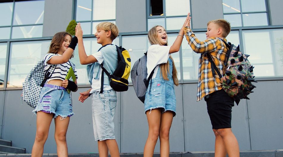 Junge Schülerinnen und Schüler klatschen vor einer Schule miteinander ab.