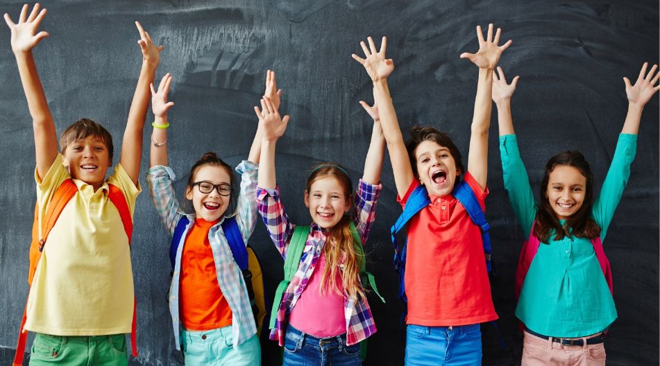 Lachende Kinder freuen sich und strecken die Hände vor einer Tafel in die Luft.