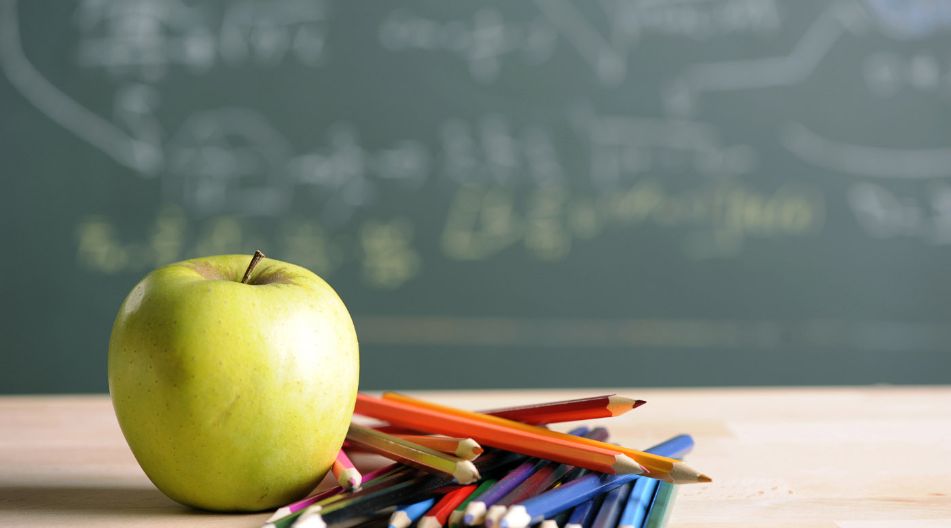 Ein grüner Apfel liegt neben einem Haufen bunter Bleistifte auf einem Holztisch. Im Hintergrund ist eine Schultafel mit unscharfen Zeichnungen und Formeln zu sehen.