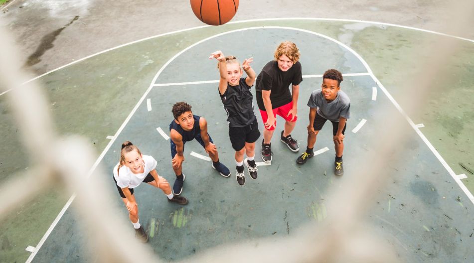 Kinder werfen einen Basketball in Richtung des Korbes. Die Kamera hängt an dem Korb und nimmt das Foto aus der Perspektive des Ringes auf.