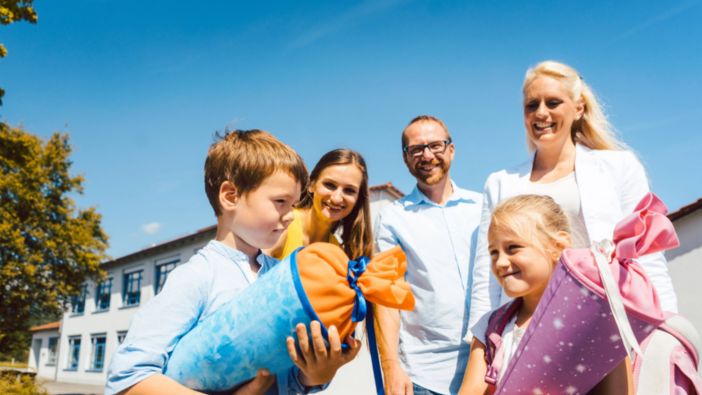 Zwei Kinder mit bunten Schultüten stehen vor einer Schule, begleitet von drei lächelnden Erwachsenen bei strahlendem Sonnenschein.