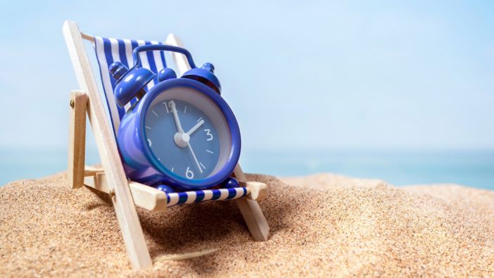 Ein blauer Wecker sitzt auf einer gestreiften Mini-Liege im Sand mit Blick auf das Meer, symbolisierend Entspannung und Urlaubszeit.