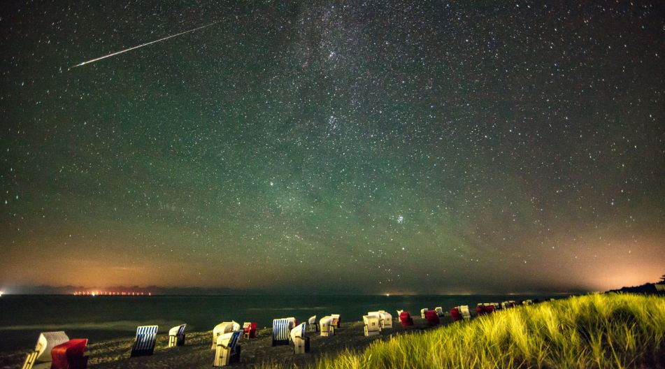 Ein Sternenhimmel über dem Meer. Am Strand stehen zahlreiche Strandkörbe, die durch den grünlich schimmernden Nachthimmel erleuchtet werden.