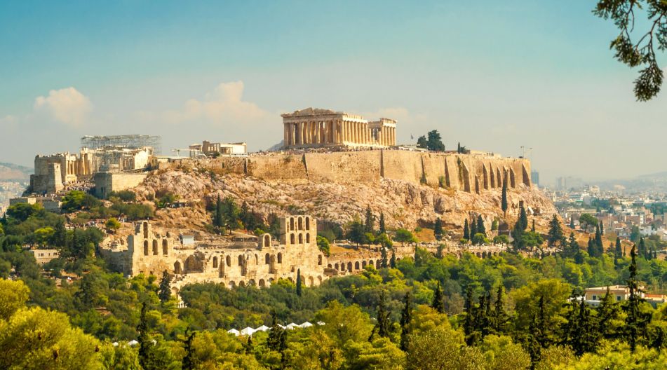 Blick auf die Akropolis von Athen, ein berühmtes antikes Bauwerk mit dem Parthenon an der Spitze eines felsigen Hügels, umgeben von üppigem Grün und der Stadt im Hintergrund.