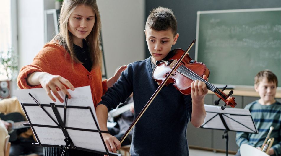 Eine Musiklehrerin hilft einem Jungen beim Geigenspiel, indem sie das Notenblatt richtet. Der Junge hält die Geige konzentriert, im Hintergrund sind weitere Kinder mit Musikinstrumenten in einem Klassenraum zu sehen. Eine Musiklehrerin hilft einem Jungen beim Geigenspiel, indem sie das Notenblatt richtet. Der Junge hält die Geige konzentriert, im Hintergrund sind weitere Kinder mit Musikinstrumenten in einem Klassenraum zu sehen.