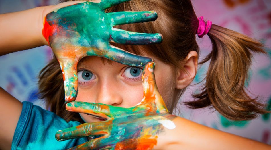 Ein Kind mit blauen Augen und braunen Zöpfen hält bemalte Hände vors Gesicht und formt mit den Fingern einen Rahmen, durch den es schaut. Die Hände sind bunt mit Farben bedeckt, im Hintergrund sind Handabdrücke an der Wand zu sehen. Ein Kind mit blauen Augen und braunen Zöpfen hält bemalte Hände vors Gesicht und formt mit den Fingern einen Rahmen, durch den es schaut. Die Hände sind bunt mit Farben bedeckt, im Hintergrund sind Handabdrücke an der Wand zu sehen.