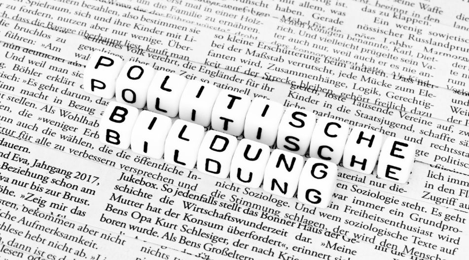 Buchstabenwürfel bilden die Worte "POLITISCHE BILDUNG" und liegen auf einer Zeitungsseite mit schwarzem gedrucktem Text.