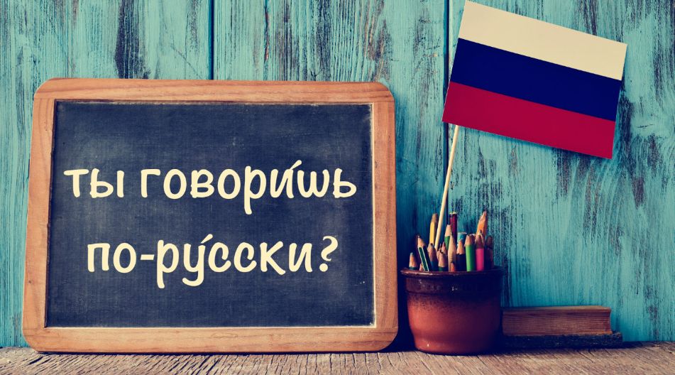 Eine Tafel mit der Aufschrift „Ты говоришь по-русски?“ vor einer blauen Holzwand, daneben ein Topf mit Buntstiften und eine kleine russische Flagge.