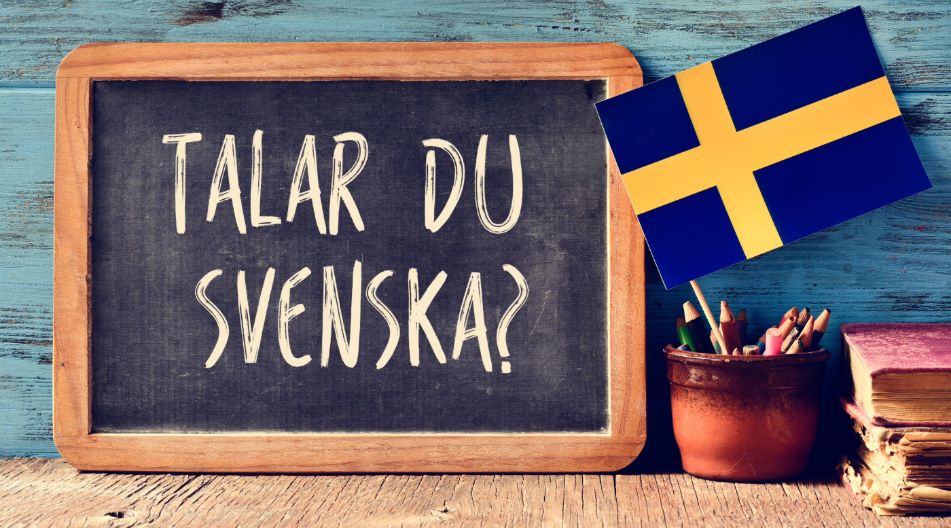 Eine Tafel mit der Aufschrift „Talar du svenska?“ vor einer blauen Holzwand, daneben ein Topf mit Buntstiften und eine kleine schwedische Flagge.