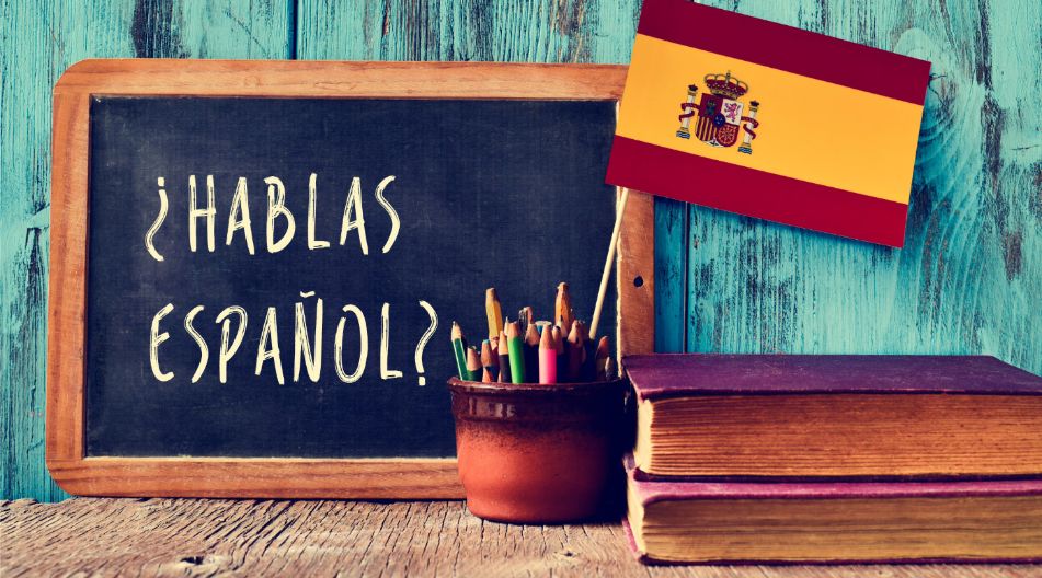 Ein Bild mit einer Schiefertafel, auf der '¿Hablas español?' steht, einer spanischen Flagge, einem Topf mit bunten Stiften und zwei Büchern, symbolisiert das Lernen und Sprechen der spanischen Sprache.